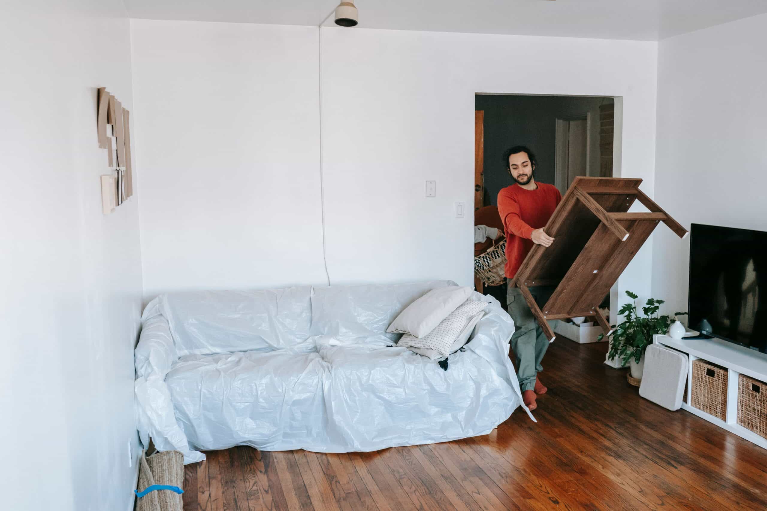 Man lifting a wooden table in a bright living room with wrapped sofa, preparing for a move.