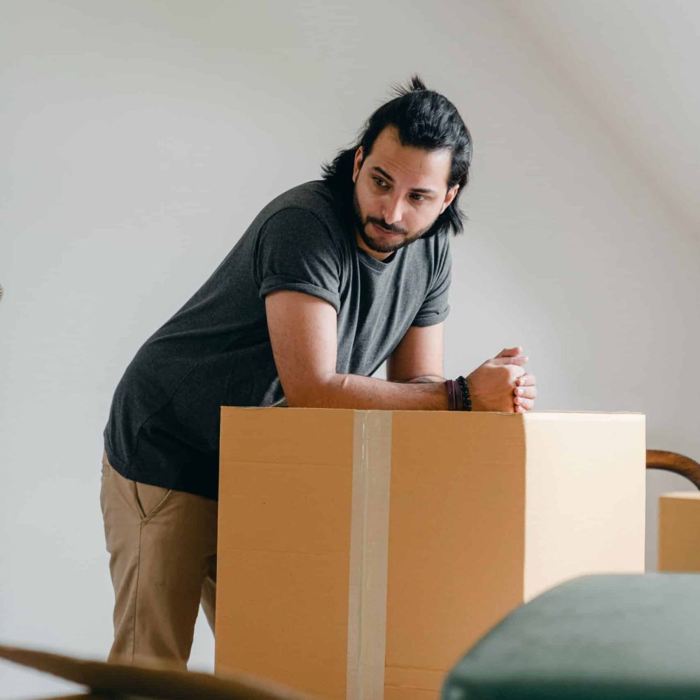 Pensive bearded male in casual clothes leaning on large carton box while moving to new house and looking away