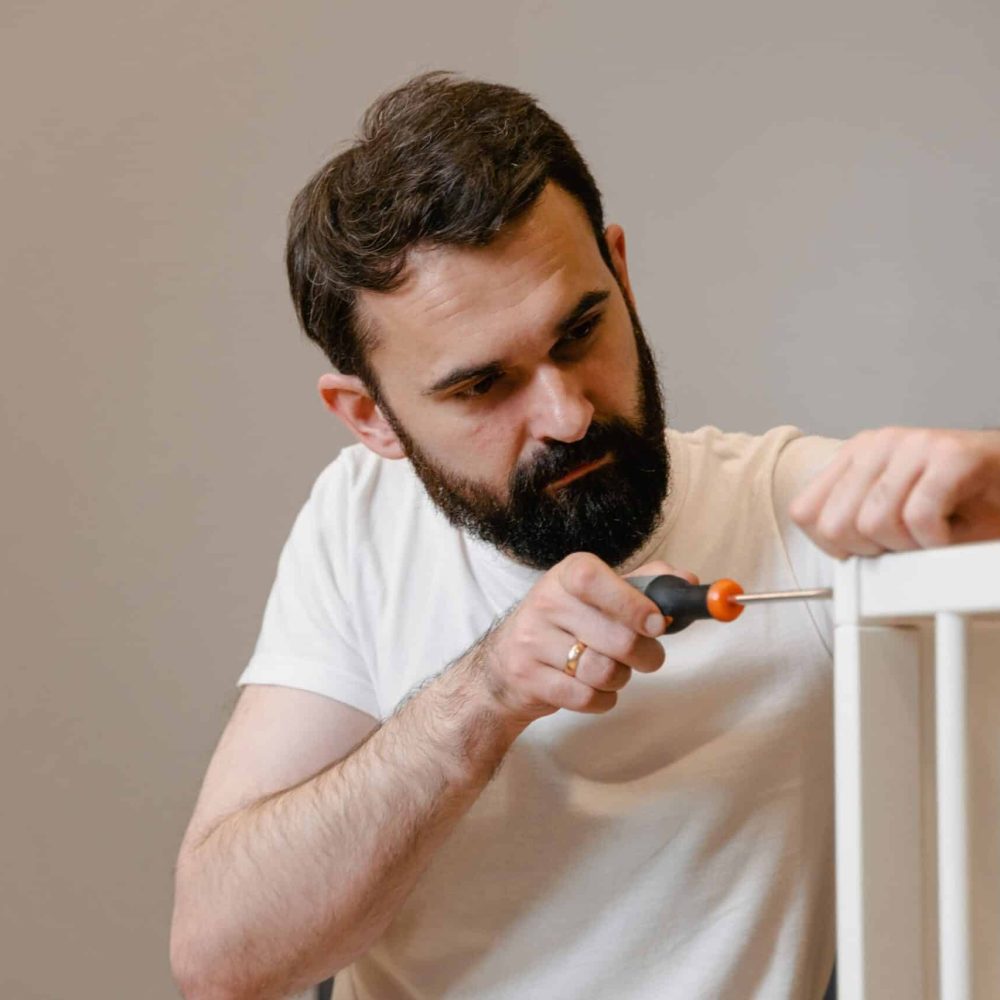A man focusing closely on assembling a baby crib with a screwdriver in a well-lit room.